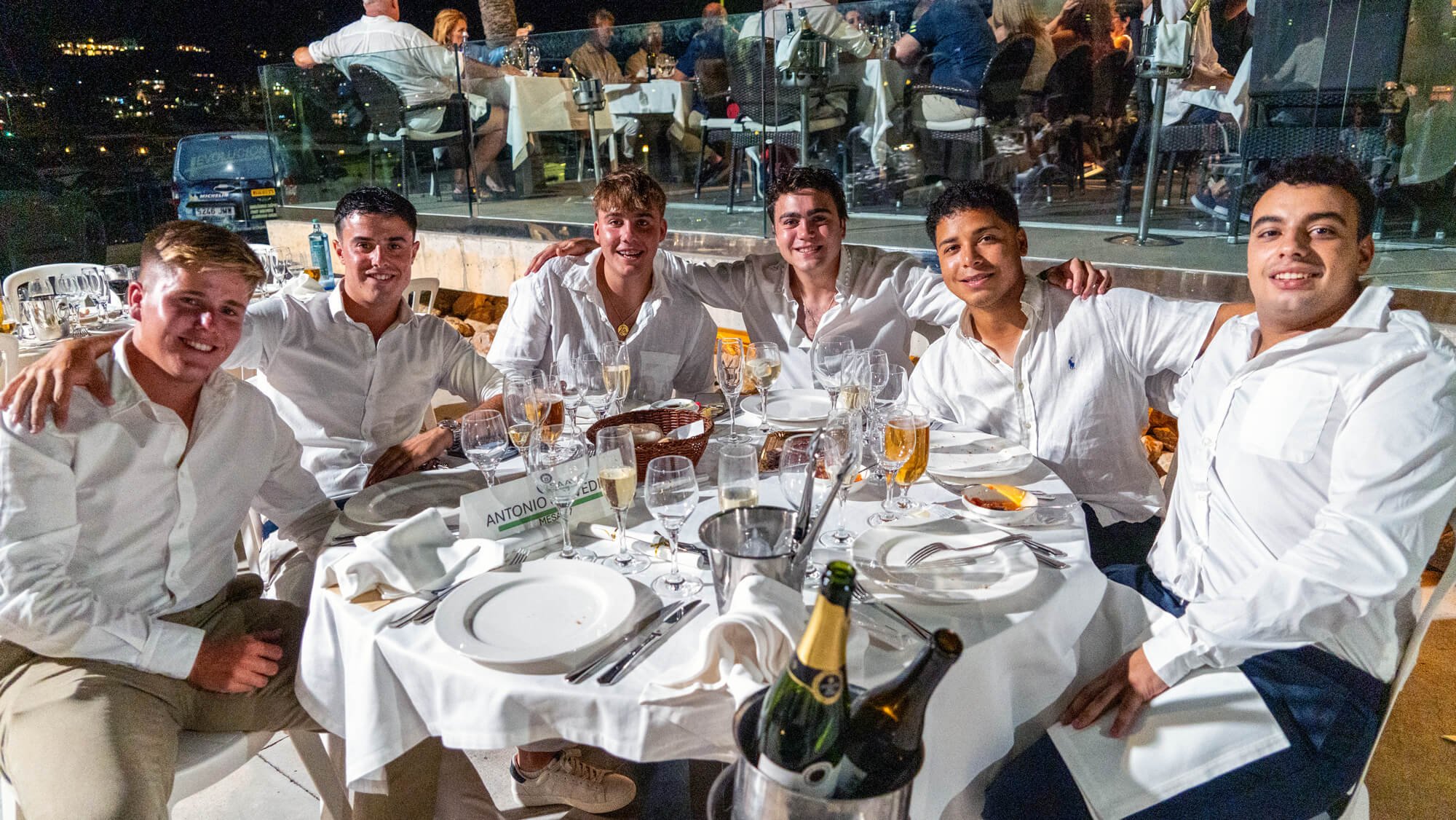 Grupo de seis jóvenes en una cena formal al aire libre por la noche, con mesas arregladas con cristalería y comida, en un ambiente elegante con vista nocturna en el fondo.