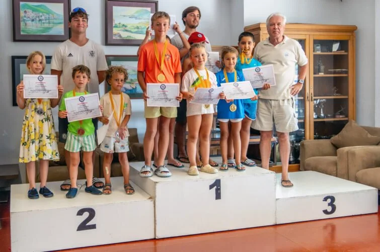 Niños en un podio recibiendo premios y certificados, con un adulto a la derecha, en una sala con pinturas en las paredes y un mueble de madera en el fondo.