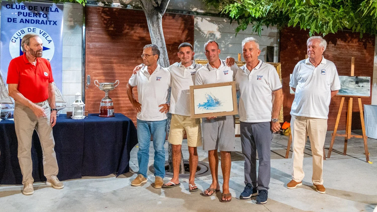 Grupo de siete personas en una ceremonia de premiación en un evento náutico, con un cartel que dice 'Club de Vela Puerto de Andratx' al fondo, algunos sosteniendo un cuadro y un trofeo, en un ambiente al aire libre con árboles y obras de arte.