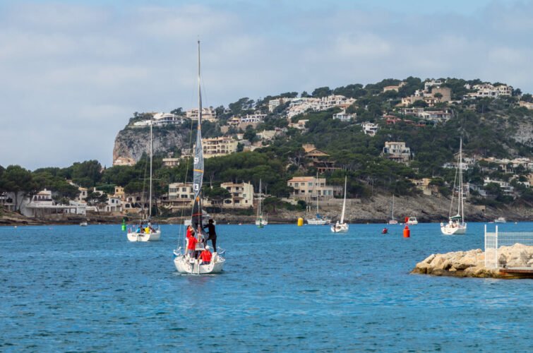 Barco de vela con personas navegando en aguas tranquilas frente a una costa con casas y colinas verdes.