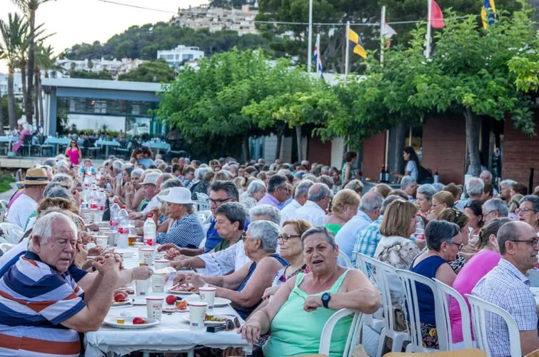 Larga mesa al aire libre con muchas personas mayores sentadas, comiendo y conversando en una reunión o evento social. Hay árboles, banderas y edificios en el fondo.