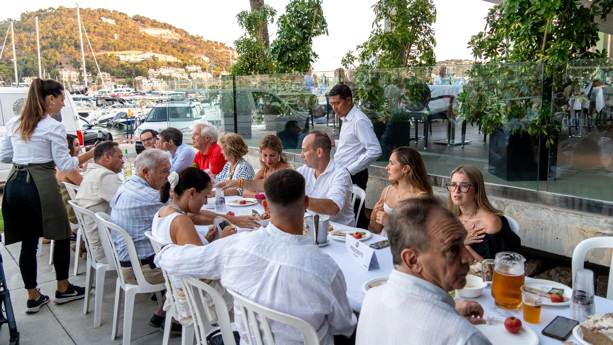 Personas en una comida al aire libre en un restaurante con vista al mar, con yates y montañas de fondo.
