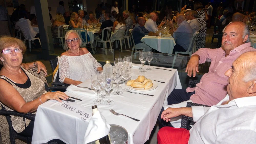 Grupo de cinco personas sentadas en una mesa en un evento o cena, con muchas mesas y personas al fondo.