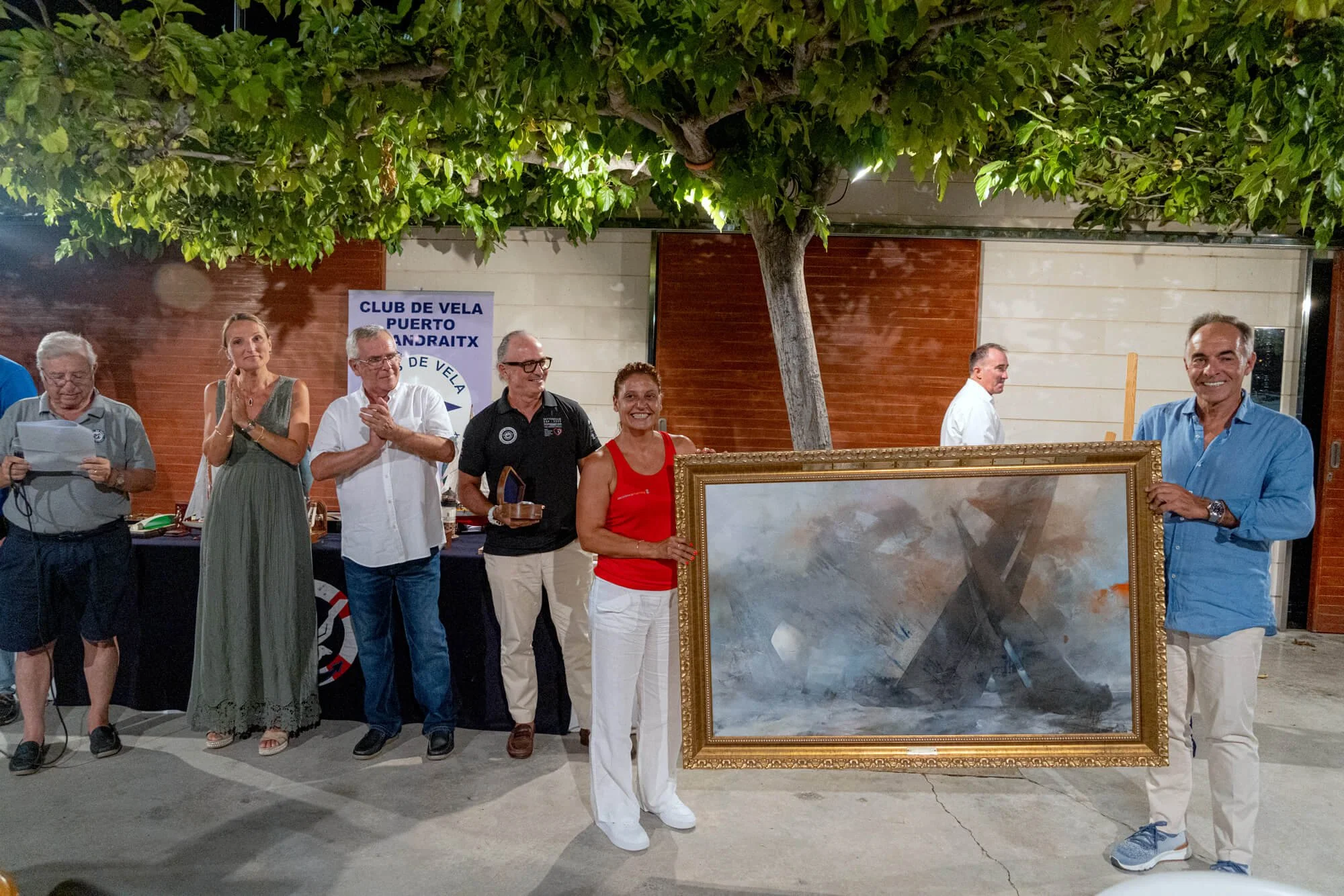 Grupo de personas en una celebración, con un árbol grande en el centro y un cartel que dice 'Club de Vela Puerto Andraitx', una mujer sostiene un cuadro grande con marco dorado, y algunas personas están sonriendo y aplaudiendo.