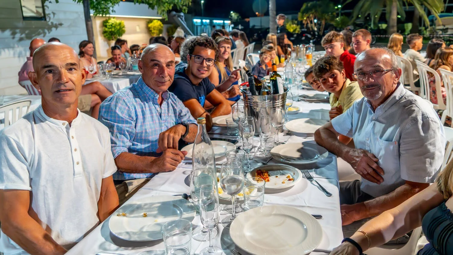 Grupo de personas celebrando en una comida al aire libre por la noche, con comida y bebidas sobre la mesa.