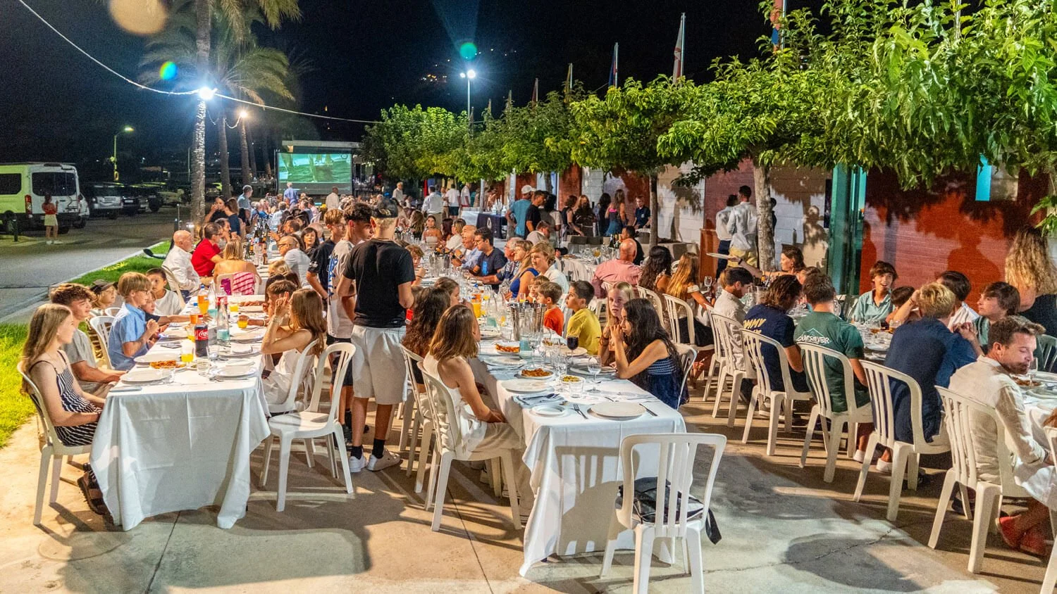 Cena al aire libre en la noche con muchas personas en mesas blancas, comiendo y disfrutando, rodeadas de árboles y luces, en un evento social.