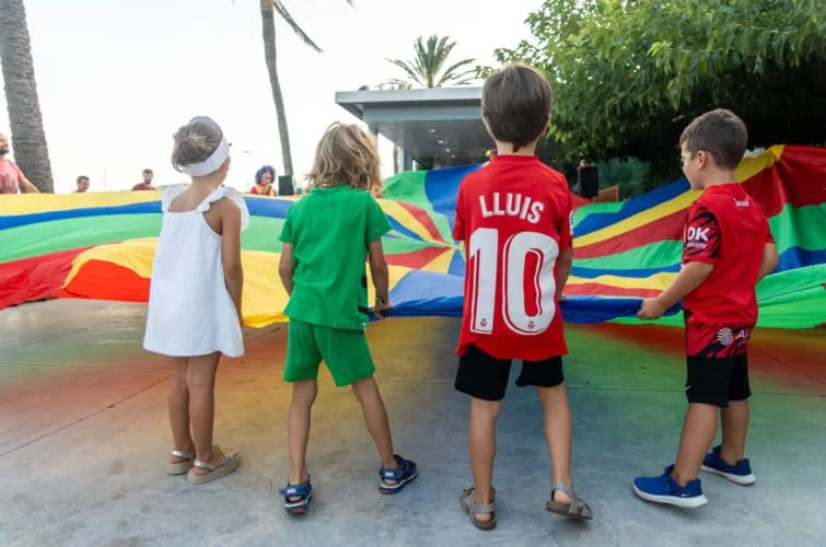 Cuatro niños jugando con un paracaídas multicolor en una zona al aire libre.