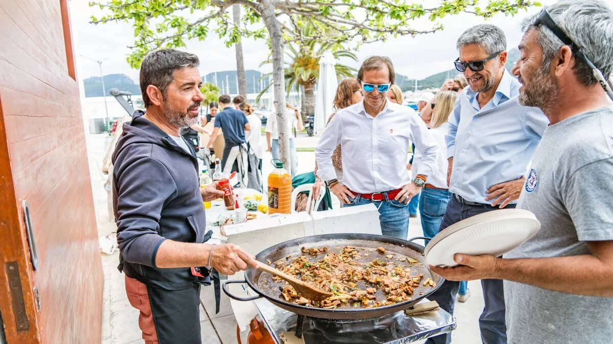 Hombre cocinando alimentos en una parrilla para un grupo de personas en una reunión al aire libre cerca del mar con árboles y montañas al fondo.