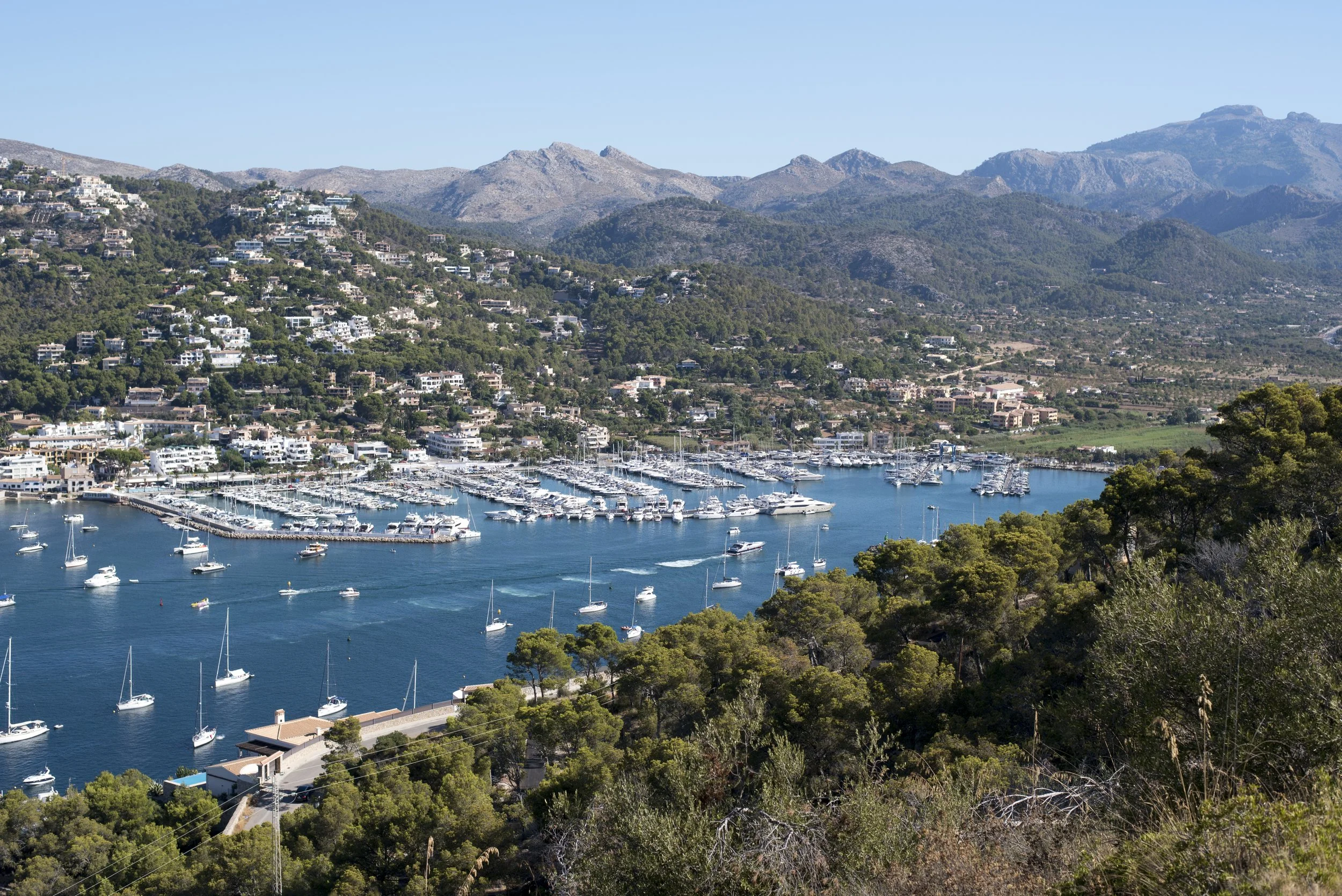 Vista panorámica de un puerto con muchas embarcaciones, rodeado de colinas con casas y montañas en el fondo bajo un cielo despejado.