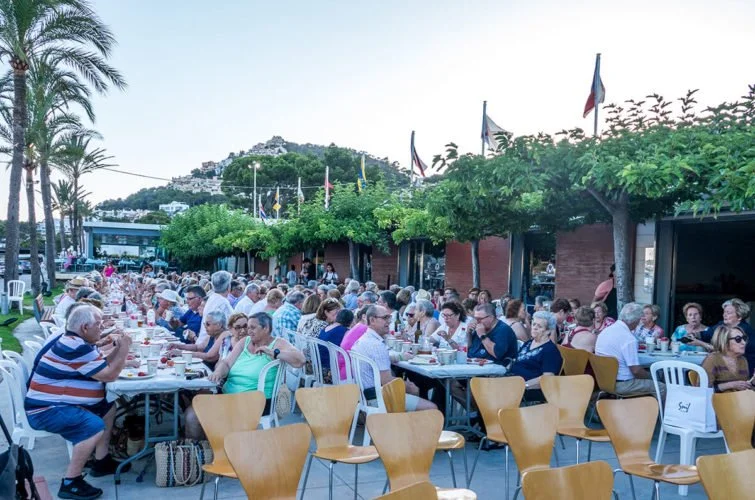 Muchas personas sentadas en mesas al aire libre disfrutando una comida o reunión en un entorno con vegetación y banderas.