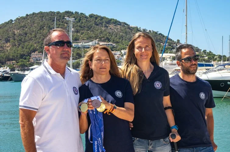 Cuatro personas posando junto a un muelle con barcos, en un día soleado, con colinas y edificios al fondo, y una de ellas sosteniendo medallas y medallones.