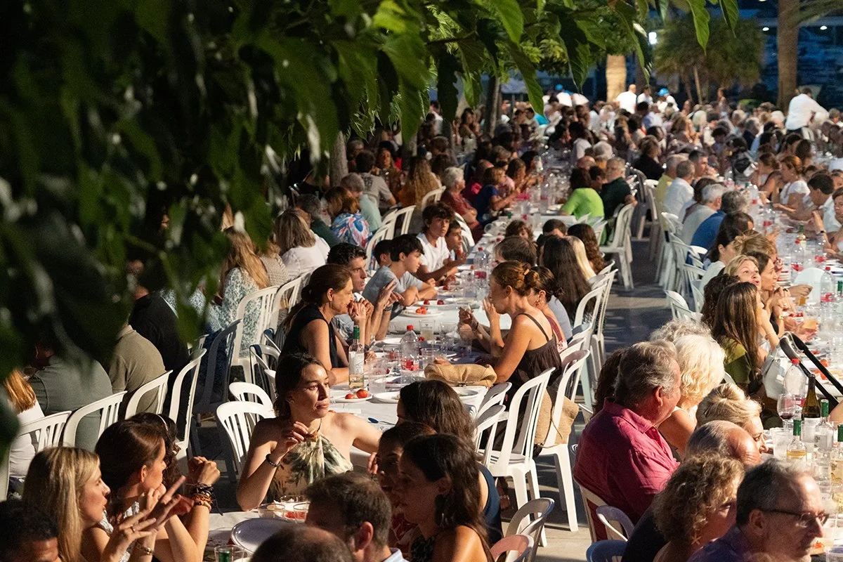 Amplio banquete al aire libre con muchas personas sentadas en mesas largas, rodeadas de árboles, en una fiesta por la tarde o noche.