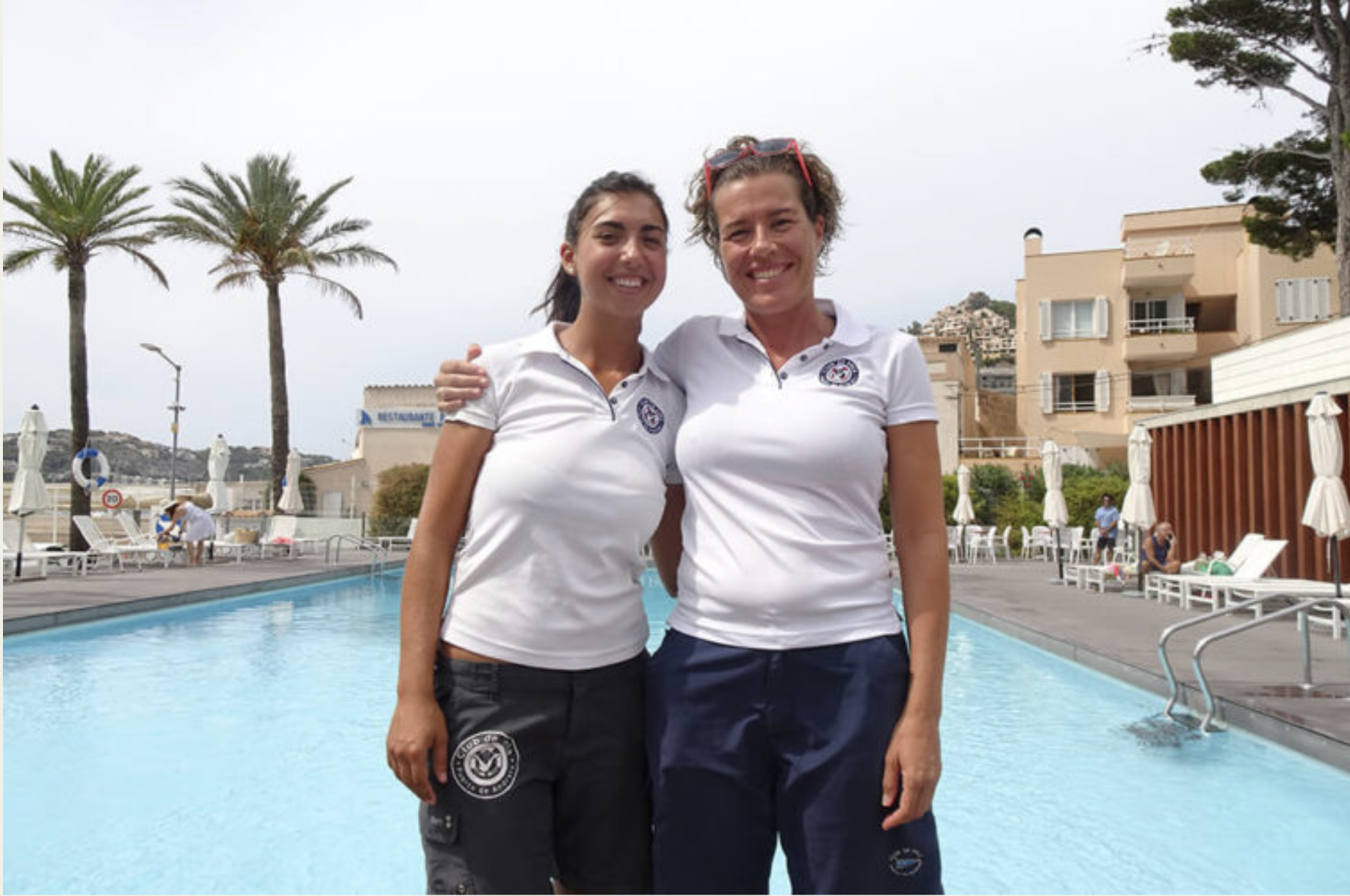 Dos mujeres con uniformes deportivos de pie junto a la piscina, sonriendo, en un área de recreo con sillas y sombrillas blancas, y edificios residenciales en el fondo.