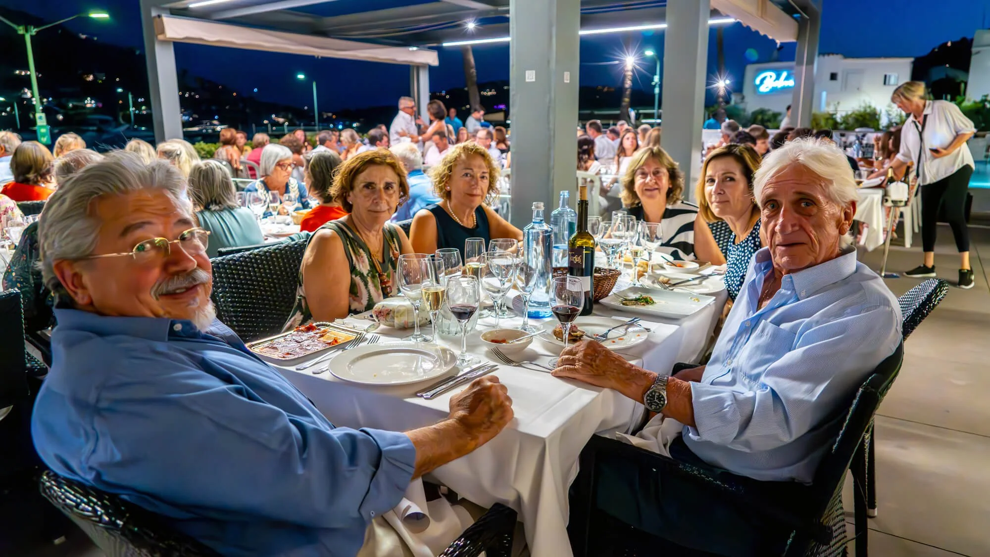 Grupo de personas mayores sentadas en una mesa de restaurante al aire libre en la noche, disfrutando de una comida y bebida, con muchas personas en el fondo y luces en el entorno.