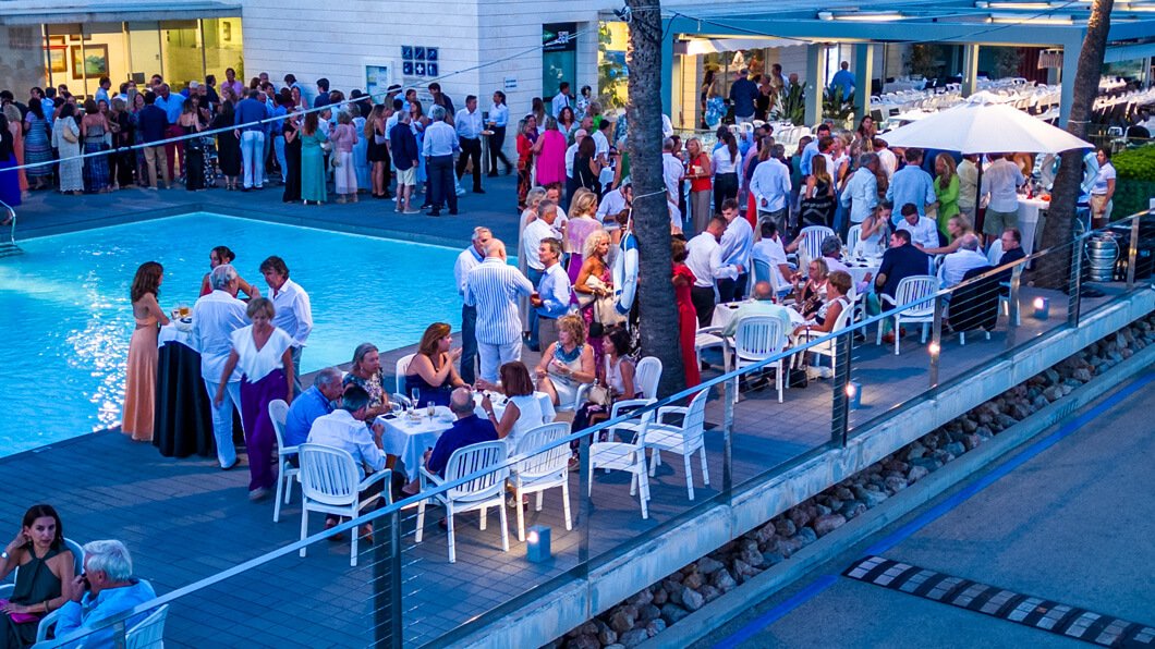 Personas en una fiesta o evento social al aire libre, con comida y bebidas, junto a una piscina, en un ambiente elegante y formal.