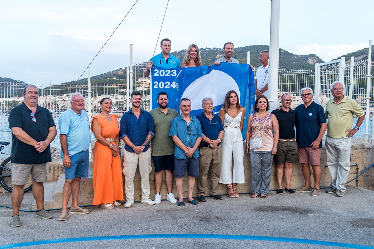 Grupo de personas de pie frente a una reja, sosteniendo una bandera con los años 2023/2024 y una luna blanca, en un entorno con montañas en el fondo.