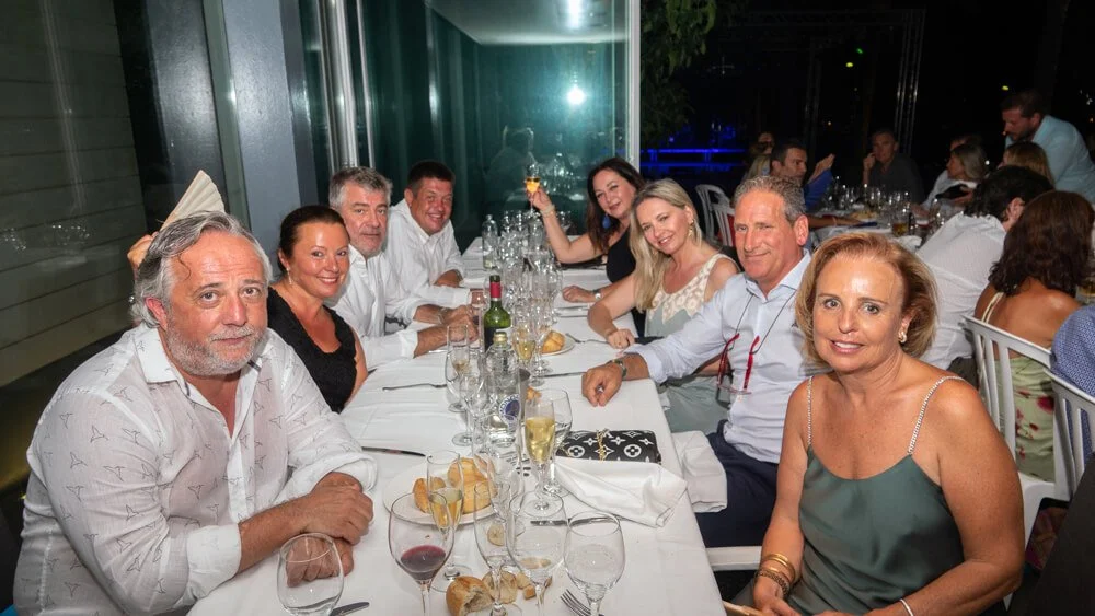 Un grupo de adultos disfrutando una cena en un restaurante, sentados alrededor de una mesa con comida, vino y copas, en un ambiente festivo y alegre.