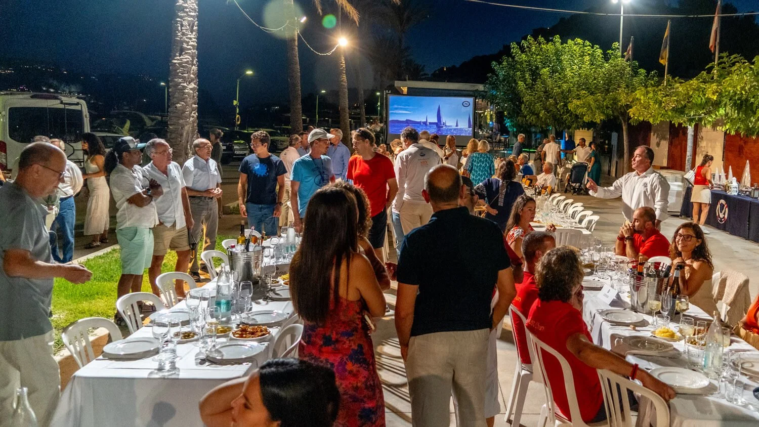 Reunión social en una terraza nocturna con mesas servidas y un grupo de personas conversando y disfrutando la noche, con una pantalla grande en el fondo y palmeras alrededor.