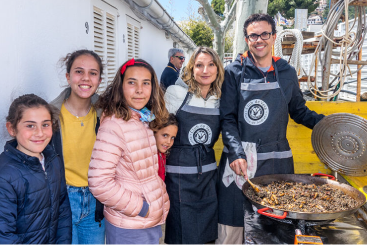 Grupo de seis personas, incluyendo niños y adultos, son sonriendo y participando en una actividad de cocina o comida en un evento al aire libre, con un foco en una sartén grande con comida.