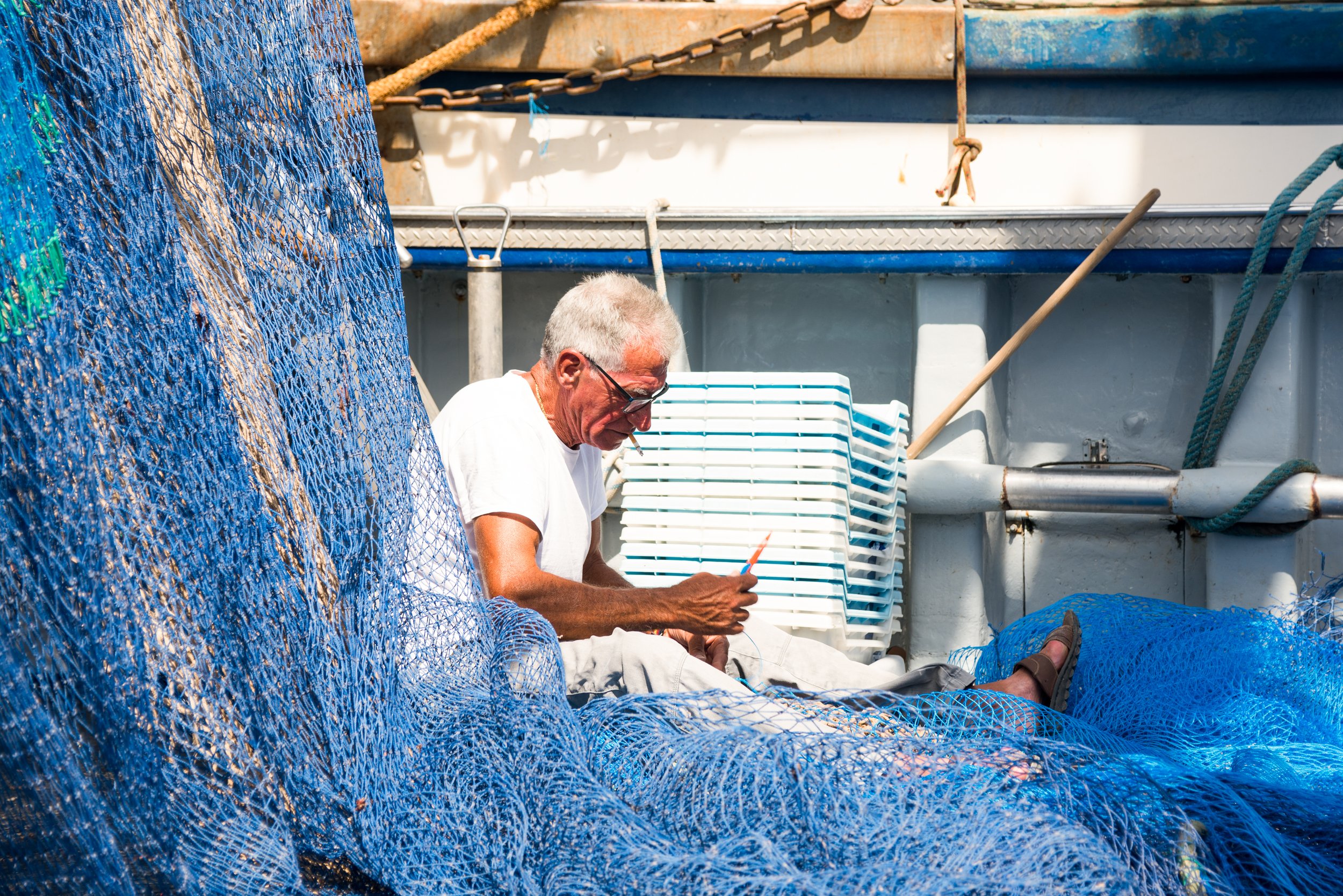 Hombre de cabello canoso con gafas, sentado en una embarcación de pesca, rodeado de redes de pesca azules y otros equipos de pesca.