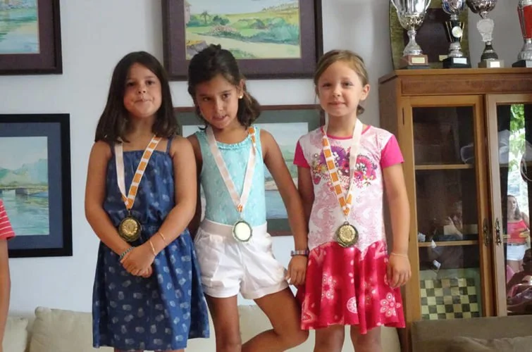 Tres niñas con medallas en un podio, celebrando en un lugar decorado con cuadros y un mueble de madera con trofeos.