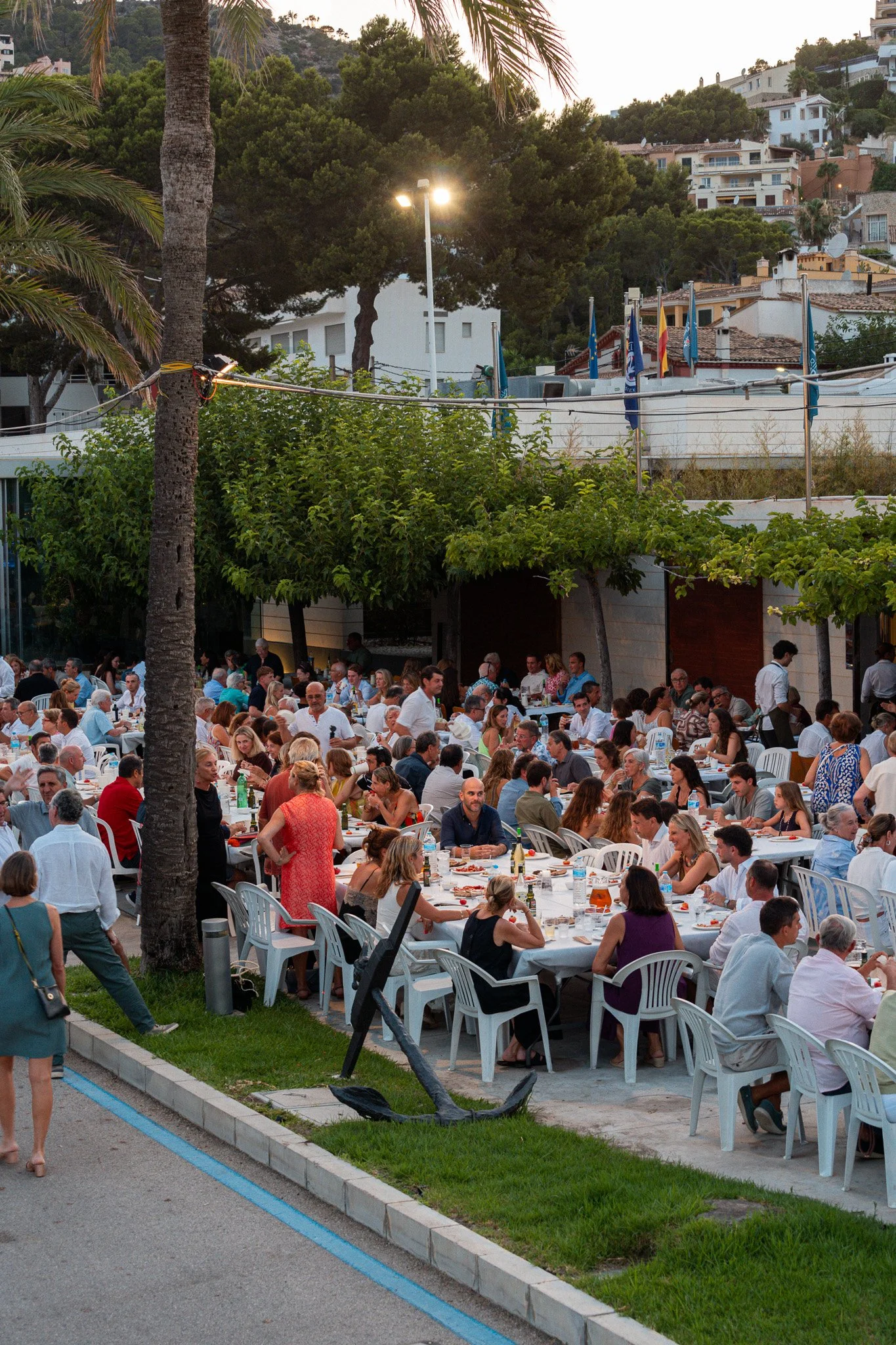 Una celebración al aire libre con muchas personas sentadas en mesas, probablemente en una comida o evento social, rodeada de árboles y un entorno urbano con casas en las colinas.