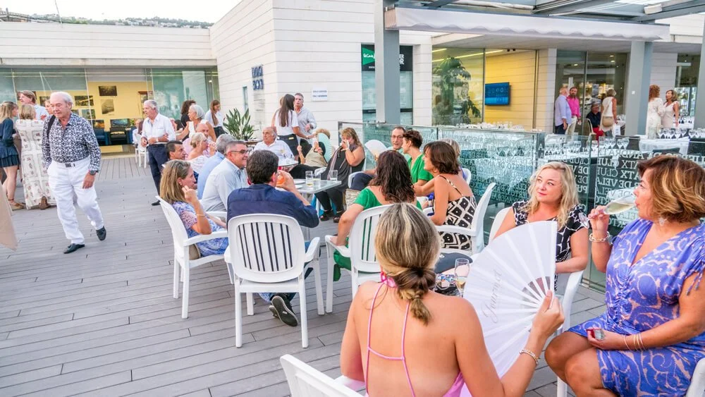 Personas charlando en una terraza al aire libre, en un ambiente social, algunas sentadas en sillas blancas, otras caminando, con una vista de un edificio moderno y decoraciones elegantes.