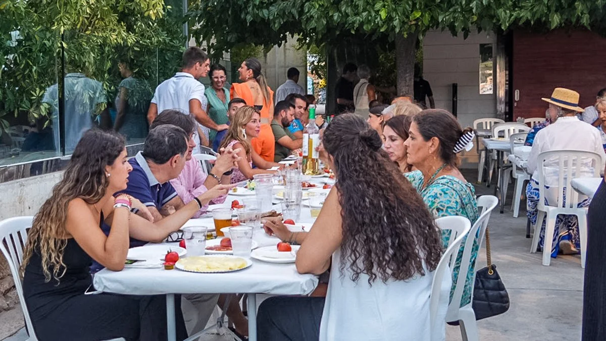 Grupo de personas sentadas en una mesa al aire libre, compartiendo comida y bebidas en un ambiente social y festivo.