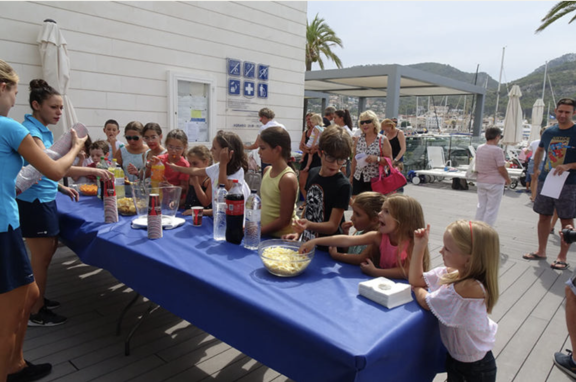 Niños y adultos en una fiesta alrededor de una mesa con alimentos en el muelle, con yates y montañas de fondo.