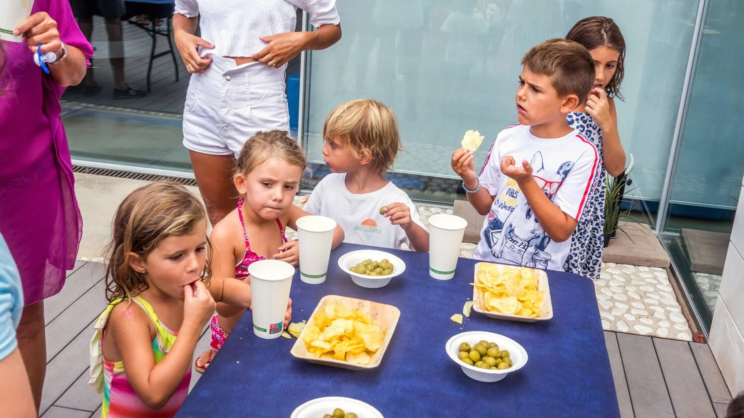 Niños pequeños en una fiesta, algunos sentados en una mesa con botellas de jugo, papas fritas y aceitunas, otros de pie y observando, en un patio con un fondo de paredes de vidrio.
