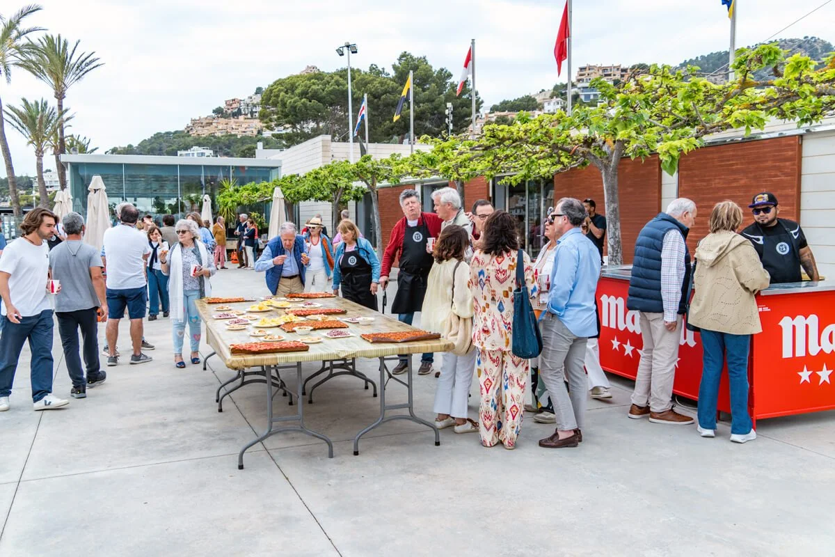 Personas comiendo en un evento con mesas y puestos de comida, palmeras y árboles, banderas y montañas al fondo.