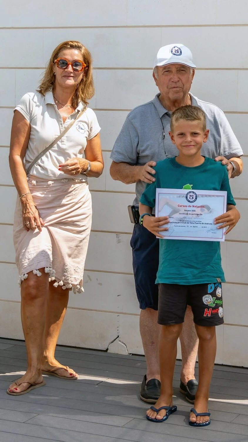 Niño con certificado de participación en cursos de natación, acompañado por dos adultos en una ceremonia, fondo de pared blanca.