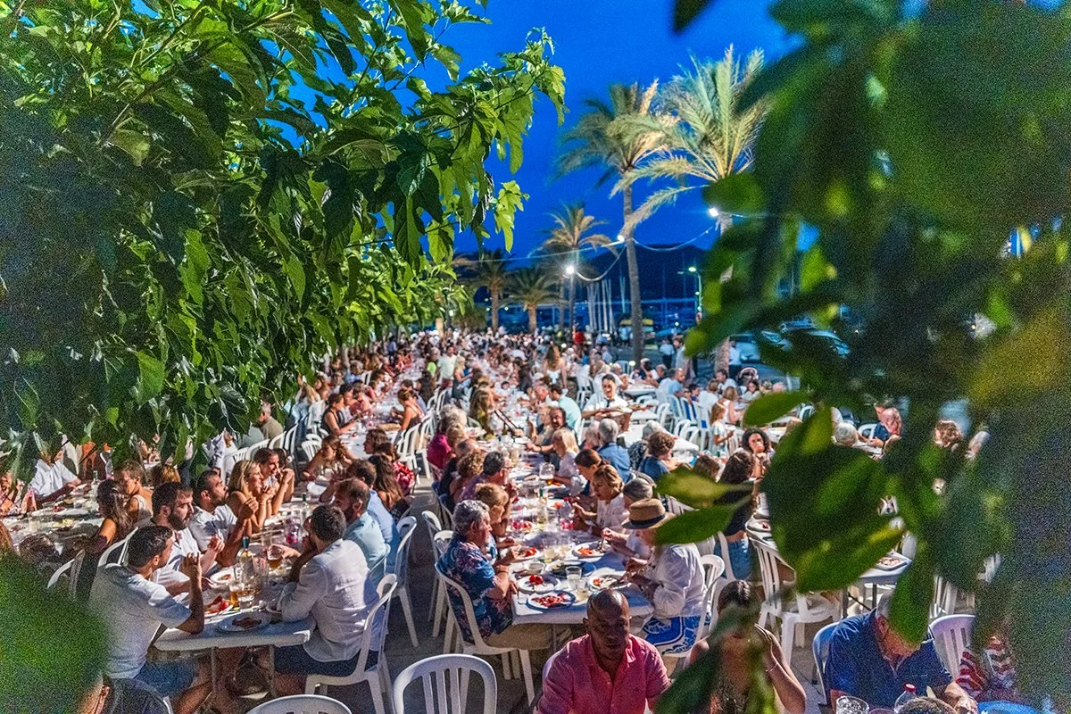 Una gran reunión al aire libre durante la noche con muchas mesas y personas comiendo y socializando, rodeadas de palmeras y árboles, en un ambiente festivo y concurrido.