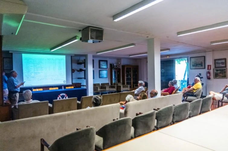 Reunión en una sala con varias personas mayores sentadas, una pantalla grande con datos, y un presentador de pie a la izquierda.