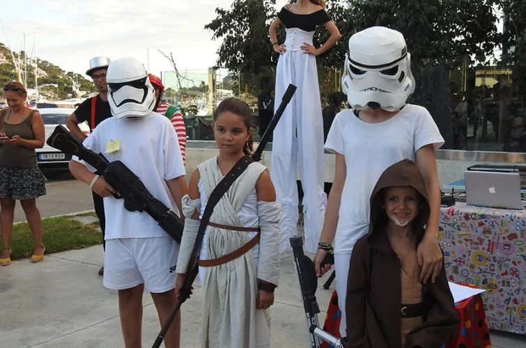 Niños disfrazados de personajes de Star Wars, algunos con cascos de stormtrooper, sosteniendo armas de juguete, en una celebración al aire libre con adultos y otros niños en el fondo.