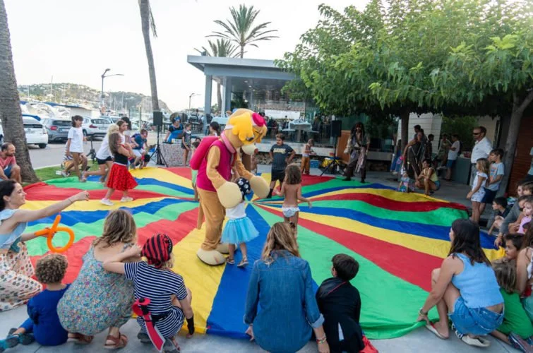 Niños y adultos jugando en una manta colorida con un personaje de mascota de payaso que lleva una camiseta rosa y pantalones beige, en un evento al aire libre cerca del mar con palmeras y puestos en el fondo.