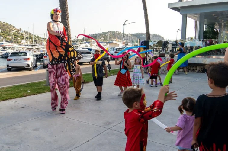 Niños disfrutando de un espectáculo con payasos y globos en un área al aire libre junto a la calle y un parque con árboles y barcos en un puerto.