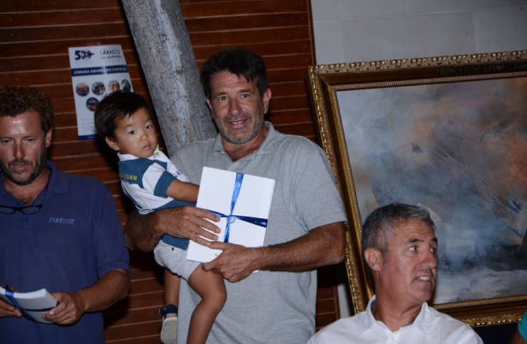 Hombre sosteniendo a un niño y un regalo con lazo azul, en un evento acompañado de otras personas, en un entorno con cuadros y cartel en la pared.