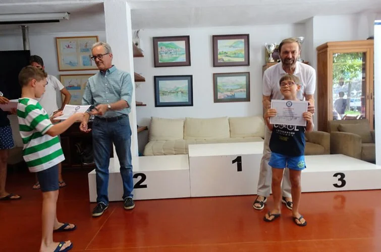 Niños y adultos en una ceremonia de premiación, con un escenario de podios numerados 1, 2 y 3, en un ambiente interior decorado con cuadros en la pared y un sofá beige.