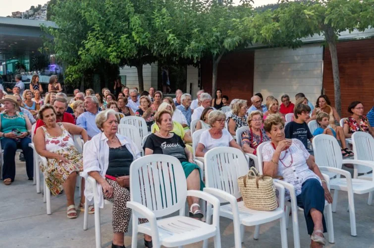 Grupo de personas sentadas en sillas blancas al aire libre, en un evento social, con árboles y un edificio de fondo.
