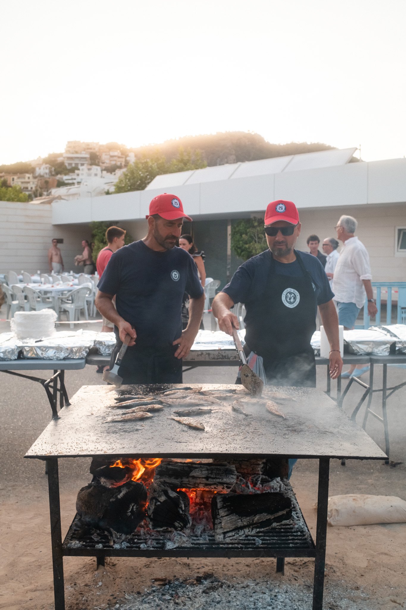 Hombres cocinando pescado en una barbacoa al aire libre, con fondo de edificios en una colina y varias personas en el área.