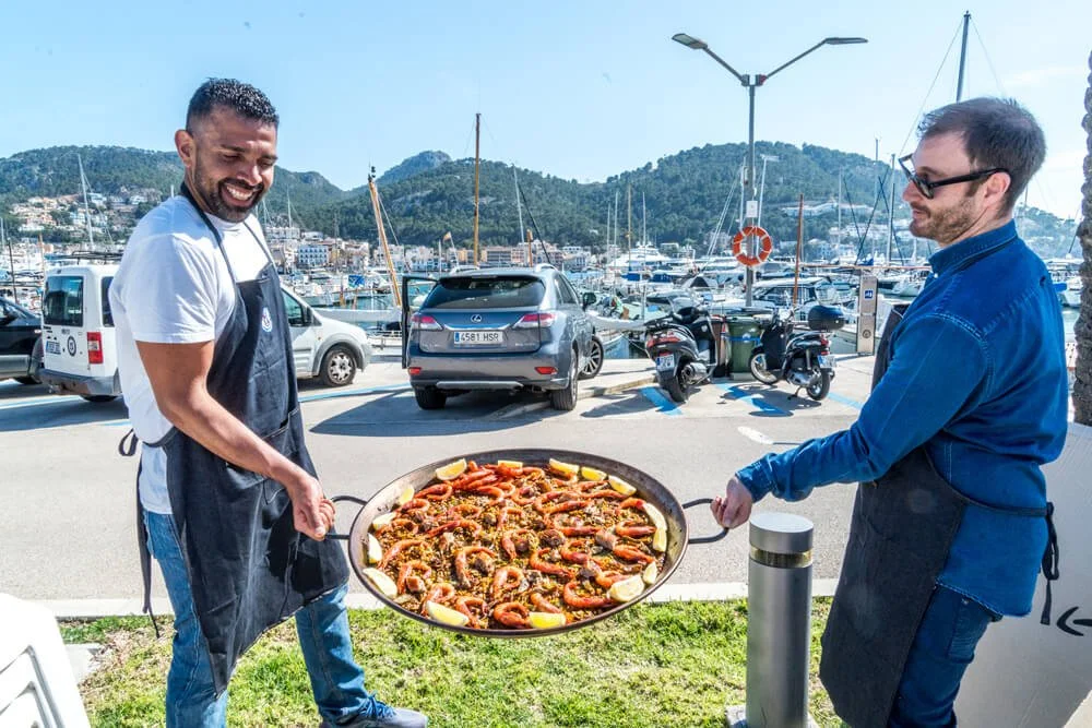 Dos hombres con delantales sosteniendo una paella con mariscos en un puerto con barcos y montañas de fondo, un día soleado.