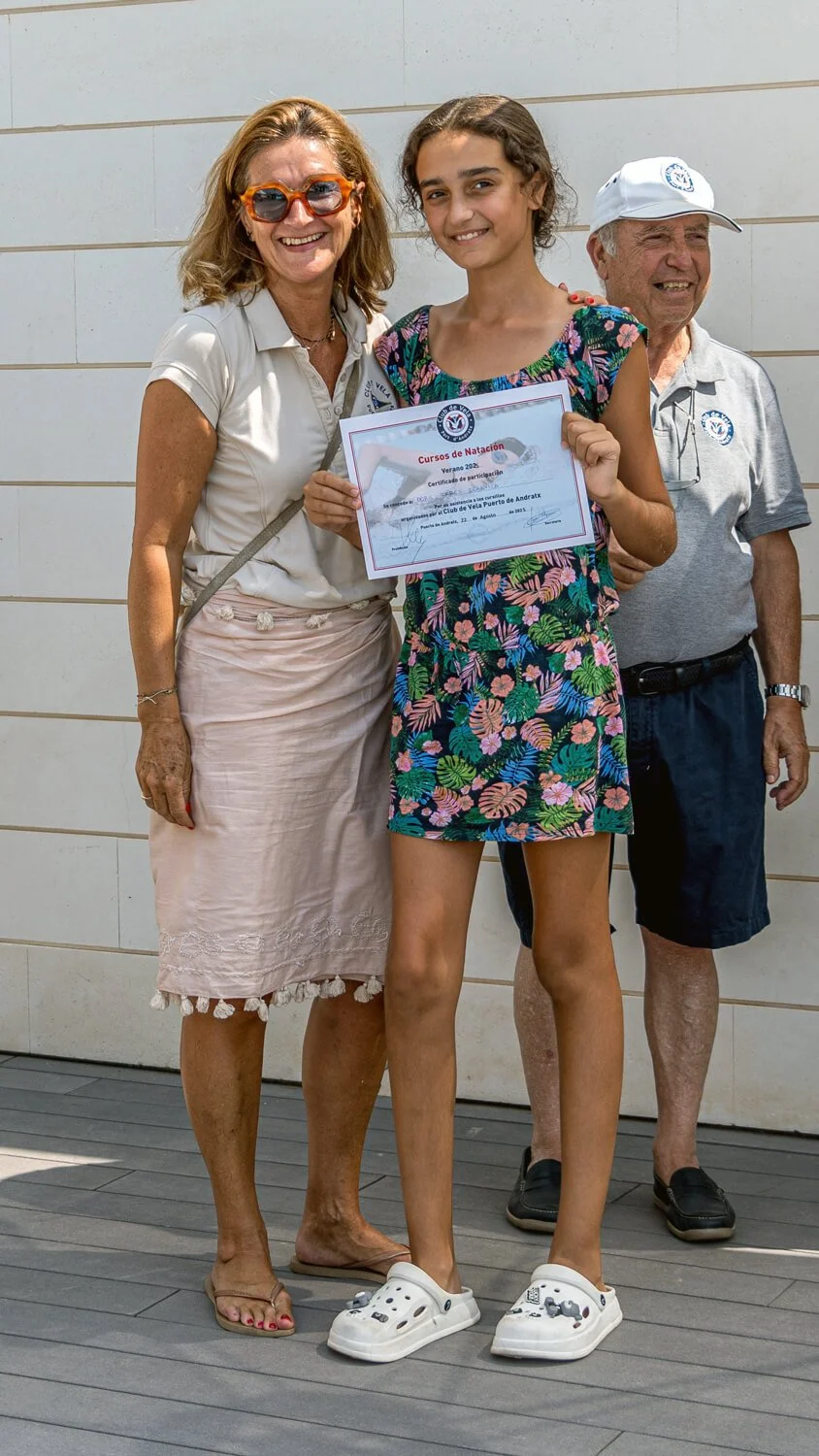 Una joven sostiene un certificado de participación en cursos de natación, acompañada de dos adultos, uno de ellos con gafas de sol y ambos con ropa casual, en un ambiente exterior.