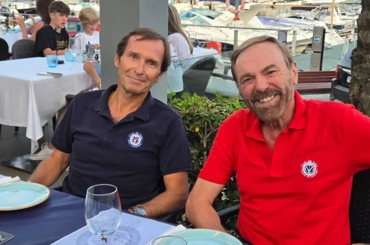 Dos hombres sentados en una mesa en un restaurante al aire libre con vista al mar y yates, uno con camiseta azul y el otro con camiseta roja, sonriendo.