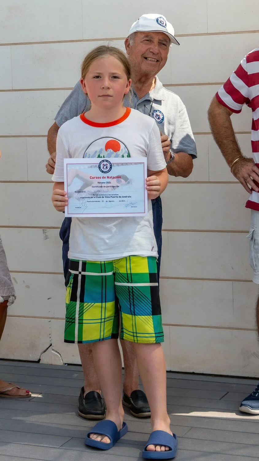 Un niño con camiseta blanca, pantalones cortos de cuadros verdes y amarillos, y sandalias azules, sostiene un certificado de participación en cursos de natación en una ceremonia. Un adulto mayor está detrás de él, sonriendo, en un fondo de pared clar