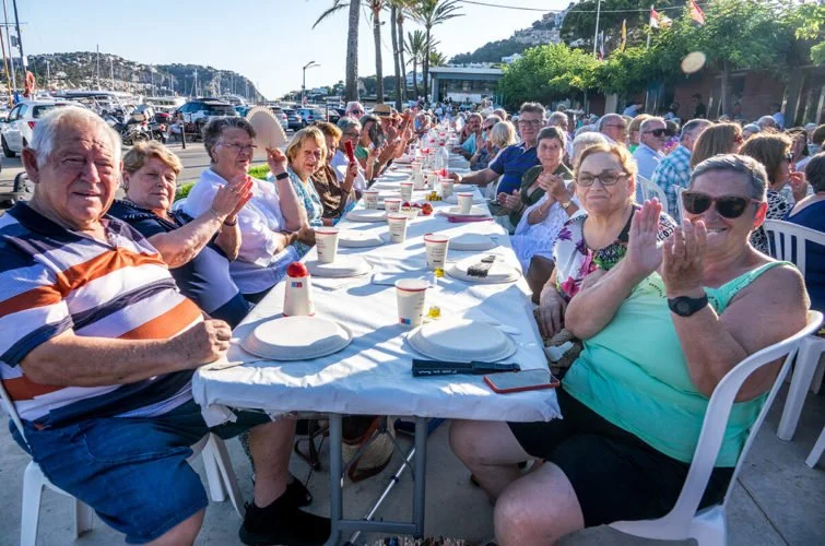 Grupo de personas mayores sentadas en una mesa larga al aire libre durante una celebración, con comida y bebidas, en un entorno marítimo con embarcaderos y árboles.