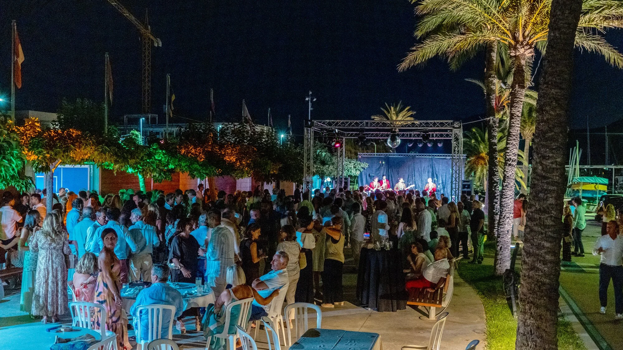 Concierto al aire libre por la noche con muchas personas disfrutando, una banda tocando en el escenario con luz de discoteca y palmeras decorativas en un entorno tropical.