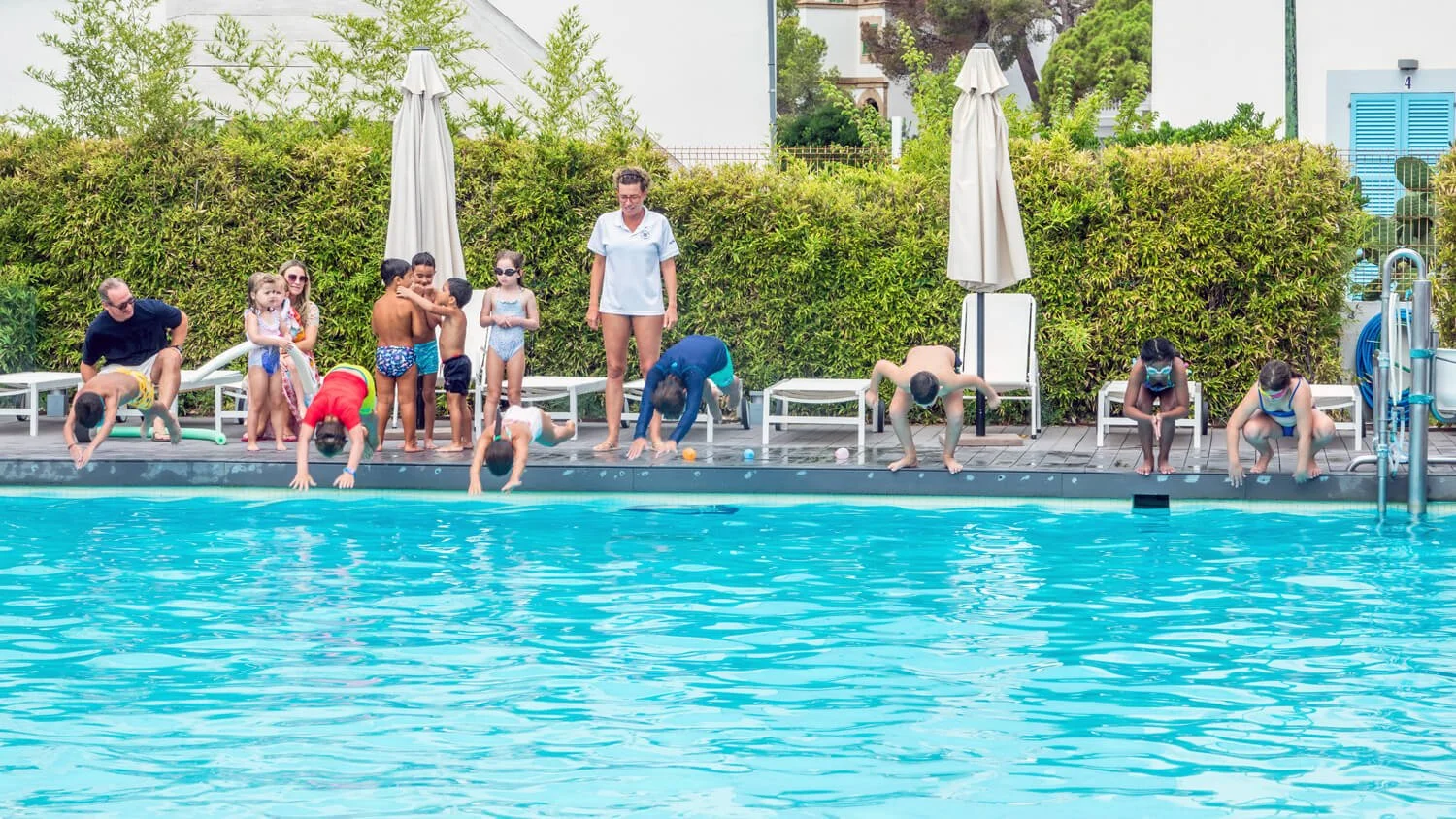 Niños haciendo clavados en la piscina, algunos adultos observando, con vegetación y sombrillas al fondo.