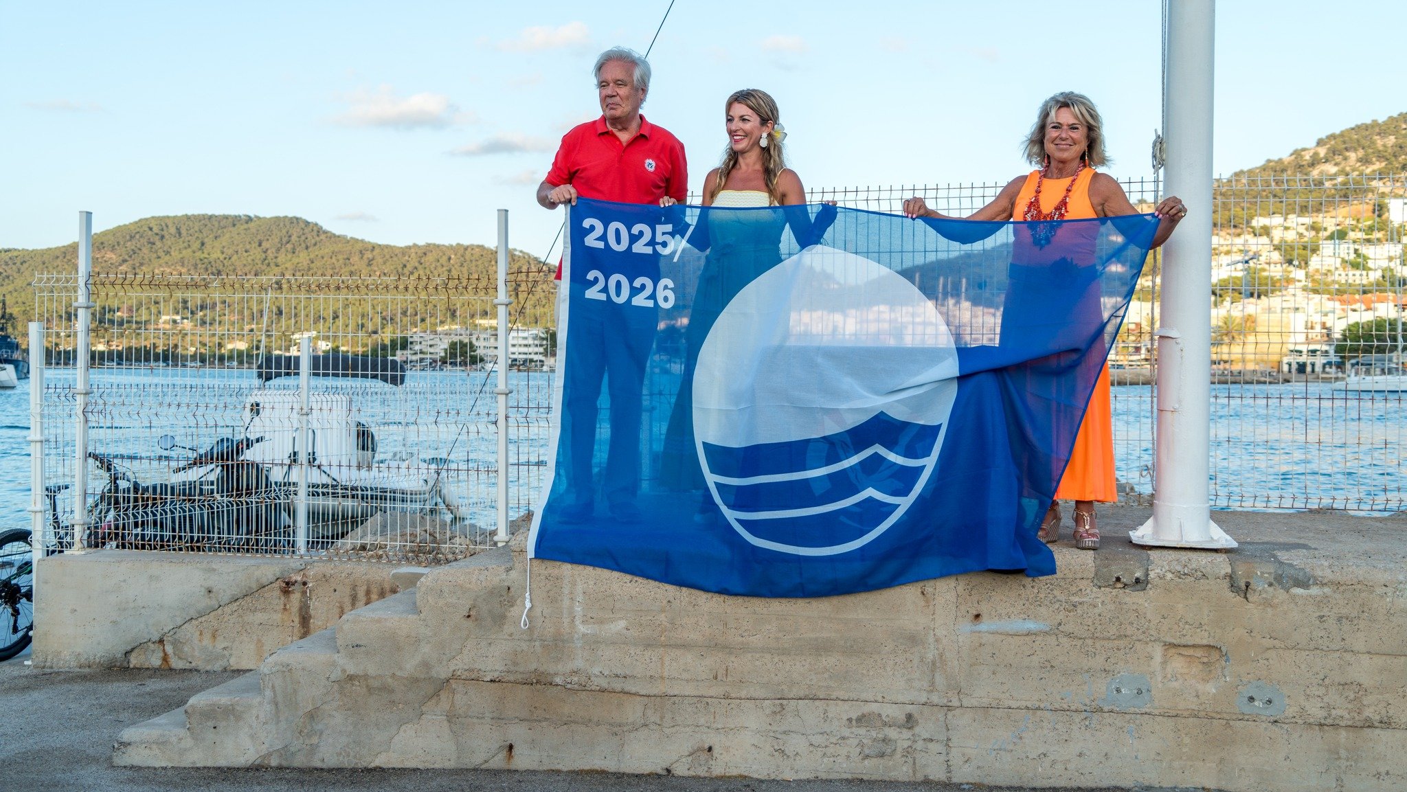 Tres personas sosteniendo una bandera azul con un logotipo y los años 2025 y 2026, en un entorno junto al agua con montañas al fondo, celebrando o participando en un evento.