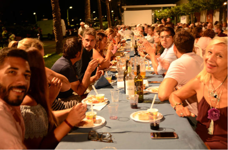 Personas sentadas en una mesa larga al aire libre, comiendo y bebiendo, en un evento social nocturno, con muchas personas en el fondo